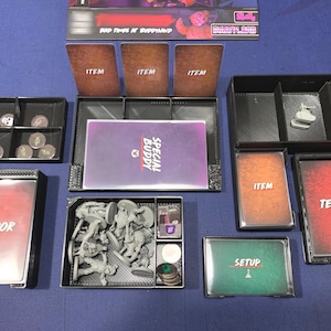 May include: A board game organizer with various components, including cards labeled "ITEM", "TERROR", and "SETUP". The game also includes miniature figures, dice, and tokens, all neatly arranged in black trays on a blue surface.