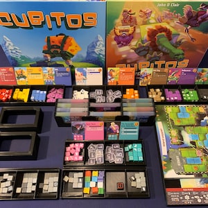 May include: A board game called "Cubitos" with colourful cubes, cards, and a game board. The game box features a cartoon character carrying a large block of cheese. The game is designed for 2-4 players.