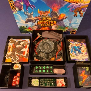 May include: A black organizer with compartments for storing game pieces for the board game "King of Monster Island". The organizer includes compartments for dice, tokens, cards, and a volcano-shaped game piece.