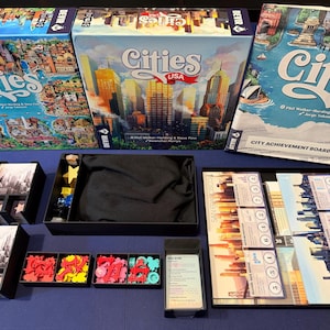 Peut inclure: Un ensemble de jeu de société comprenant trois boîtes de jeu intitulées Cities et Cities USA, un plateau de rangement noir avec un sac en tissu, des porte-cartes noirs, des récipients en plastique remplis de jetons rouges, jaunes et bleu sarcelle.