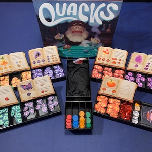 以下が含まれることがあります： 青い面に配置された、さまざまなコンポーネントを備えたボードゲーム「Quacks」。ゲームには、箱、材料トークン、プレイヤーボード、中央の大釜のイラストが含まれています。コンポーネントは黒いトレイに入っています。