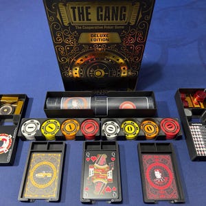Może przedstawiać: Pudełko do kooperacyjnej gry w pokera The Gang Deluxe Edition ze zdobioną grafiką w kolorze czarnym i złotym. Zawiera czarne plastikowe tacki ze stosami białych, żółtych, pomarańczowych i czerwonych żetonów, talię kart oraz kartę z rankingiem układów.