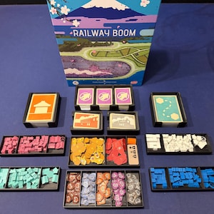 May include: The Railway Boom board game box sits above several black plastic trays containing various game components, including stacks of cards with orange, purple, and teal backs, and small wooden tokens in pink, yellow, red, white, teal, blue, and brown.