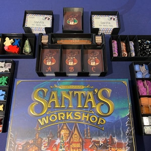 Op de afbeelding: Een zwarte organizer voor het bordspel "Santa's Workshop: 7th Edition". De organizer bevat vakken voor kaarten, tokens en andere spelonderdelen. Het bordspel is zichtbaar op de achtergrond.