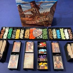 Puede incluir: Un organizador negro con múltiples compartimentos para guardar piezas de juego y cartas. El organizador está etiquetado como "Skara Brae" y presenta una ilustración colorida de una mujer sosteniendo una lanza.