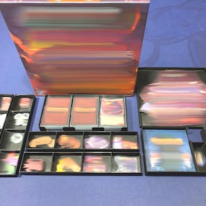 May include: A board game with various components arranged on a blue surface. The game includes a box with a colorful abstract design, multiple black trays, and stacks of cards. The trays contain game pieces and other elements.