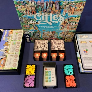 May include: A Cities board game set with a box, game board, cards, and colored tokens. The box features a colorful illustration of a city skyline. The game board has a map of a city with different colored areas. The cards are in a black tray with a red star on each side. The tokens are in small trays with different colors.