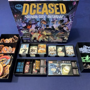 Può includere: Gioco da tavolo "DCeased: Gotham City Outbreak" con componenti. La scatola mostra Batman e altri personaggi. Il gioco include carte, gettoni, dadi e miniature, organizzati in vassoi neri.