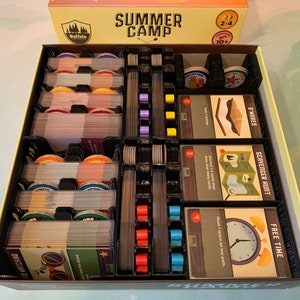 Könnte beinhalten: Ein schwarzer und grauer Spieleinsatz für das Brettspiel "Summer Camp". Der Einsatz hat Fächer für Karten, Spielsteine und andere Spielkomponenten. Der Einsatz ist so konzipiert, dass er in die Spielbox passt.