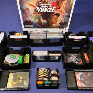Może przedstawiać: Czarne plastikowe pudełka do przechowywania z przegrodami do organizowania elementów gry. Pudełka są oznaczone "Tales to Amaze" i "Heroes vs Villain".