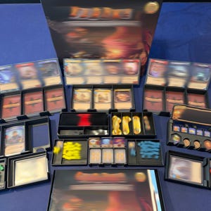 May include: A board game set featuring a large box, multiple black plastic card organizers, and various game components including decks of cards, small yellow and blue tokens, and red markers arranged on a flat surface for gameplay organization.