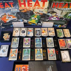 May include: A board game set with three boxes labeled "HEAT" and expansion titles. The game includes cards, miniature race cars, and other game components. The boxes feature race cars and the titles "Heavy Rain", "Pedal to the Metal", and "Tunnel Vision".