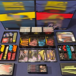 May include: A board game set featuring two large rectangular boxes with yellow and red accents, multiple black plastic organizers filled with various game components, including stacks of cards, small colored tokens, and character standees.
