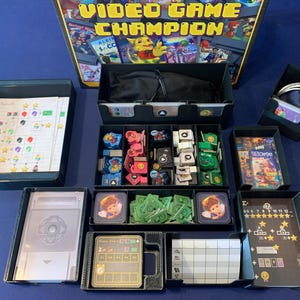 May include: A board game titled "VIDEO GAME CHAMPION" with various game components. The game includes cards, tokens, money, and a game board, all organized in black trays. The box art features colorful video game-themed graphics.