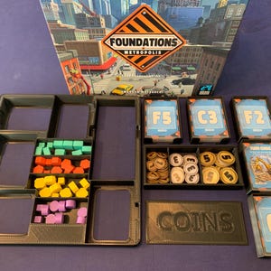 Puede incluir: Un organizador negro con múltiples compartimentos para guardar piezas de juego. El organizador tiene una etiqueta que dice "MONEDAS" e incluye compartimentos para guardar monedas, cartas y otras piezas de juego. El organizador está diseñado para el juego de mesa "Foundations of Metropolis".