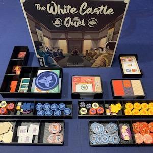 May include: The White Castle Duel board game with various game pieces, including cards, tokens, and player pieces, arranged in black trays. The game box features an illustration of a Japanese interior scene.