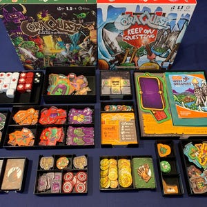 May include: Board game titled "OraQuest" with various game pieces, including dice, cards, and tokens, arranged in black trays. The game box features colorful fantasy artwork. The game is set on a blue surface.