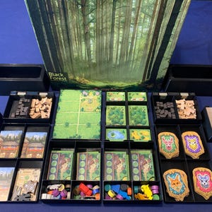 May include: A board game titled "Black Forest" with various game pieces, cards, and a game board. The game components are organized in black trays. The box art features a forest scene. The game includes wooden tokens and colorful game pieces.