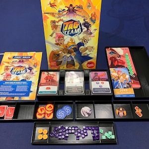 May include: A board game set, featuring a box with the title "TAG TEAM" and various game components. The set includes cards, tokens, and a rulebook, all arranged in black trays. The game's artwork features colorful illustrations of characters.