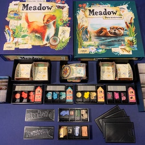 以下が含まれることがあります： 2つのボードゲームの箱、MeadowとMeadow Downstream。それぞれの箱には、トレイに整理されたゲームの部品が入っています。トレイには、カード、トークン、その他のゲームピースが入っています。箱はそれぞれ青と緑で、前面に動物のイラストが描かれています。