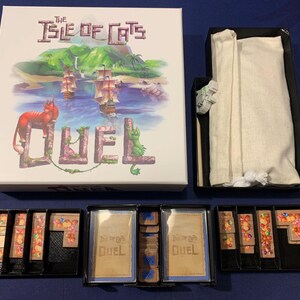 Puede incluir: Juego de mesa The Isle of Cats: Duel. La caja presenta una ilustración de una isla con barcos y gatos. El juego incluye piezas, cartas y una bolsa de almacenamiento.