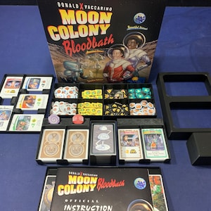 May include: The board game "Moon Colony Bloodbath" is displayed with its components. The box shows astronauts on the moon. The game includes cards, tokens, and black plastic trays. The game is set on a blue surface, with the box and components arranged.