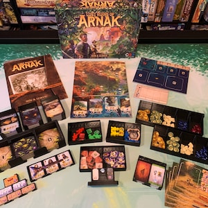 Op de afbeelding: Een spelorganisator voor het bordspel Lost Ruins of Arnak. De organisator is gemaakt van zwart plastic en heeft compartimenten voor alle onderdelen van het spel, inclusief kaarten, tokens, dobbelstenen en tegels. De organisator wordt getoond op een tafel met de doos van het spel, de regelboek en andere onderdelen.