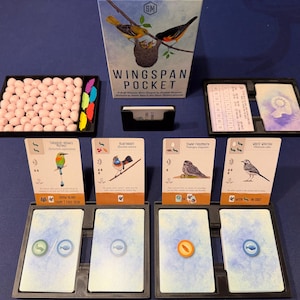 May include: Wingspan Pocket board game set featuring a game box with bird illustrations, a tray of small pink egg tokens, colorful food tokens, card holders, and several bird cards with detailed artwork and text displayed in black plastic trays.