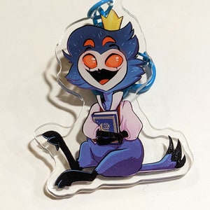 May include: A clear acrylic keychain featuring a cartoon character with blue fur, orange eyes, and a yellow crown, wearing a light pink shirt. The character is sitting and holding a blue book with a gold moon and star emblem on the cover.