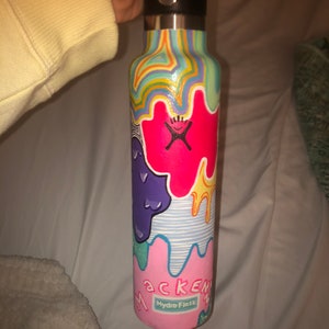 Pode incluir: Uma garrafa de água Hydro Flask com tampa preta e um design abstrato colorido. A garrafa apresenta um esquema de cores rosa, vermelho, roxo, amarelo e azul com o nome "Mackenzie" e o logotipo Hydro Flask.