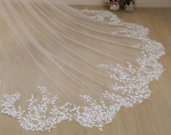 May include: A white tulle wedding veil with a delicate floral lace trim. The lace features a design of small white leaves and branches.