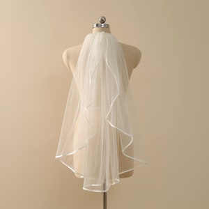 May include: A white bridal veil made from a sheer fabric. The edges of the veil are finished with a satin ribbon. The veil is displayed on a mannequin, highlighting its shape and design. A classic wedding accessory.