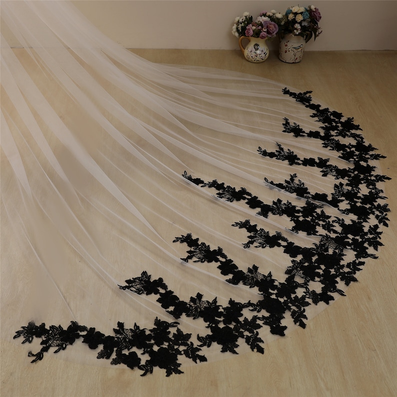May include: A white tulle wedding veil with a black floral lace trim. The veil is long and flowing, and the lace trim is delicate and intricate.