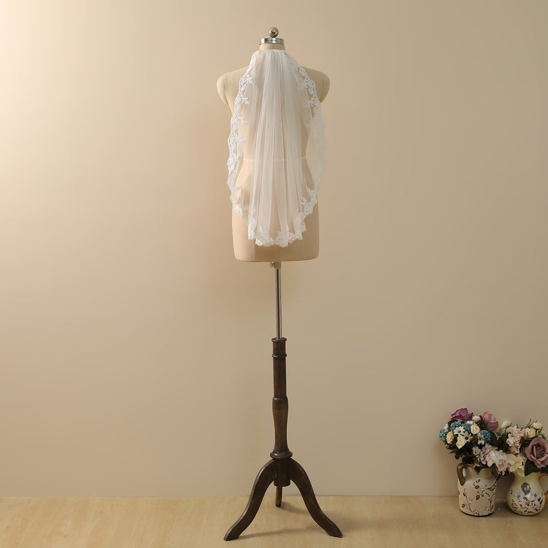May include: A white bridal veil with a delicate lace trim. The veil is displayed on a mannequin and is made of soft tulle.