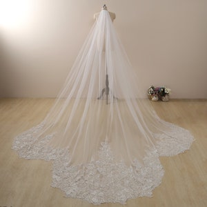 May include: A long, white tulle wedding veil with a delicate lace trim and beaded embellishments. The veil is draped over a mannequin and has a cathedral length.