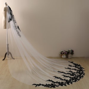 May include: A long white tulle wedding veil with a black lace trim. The veil is draped over a dress form and the lace trim is visible at the bottom of the veil.