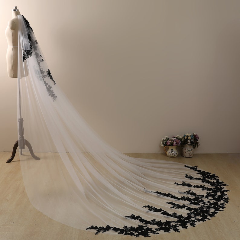 May include: A long white tulle wedding veil with a black lace trim. The veil is draped over a dress form and the lace trim is visible at the bottom of the veil.