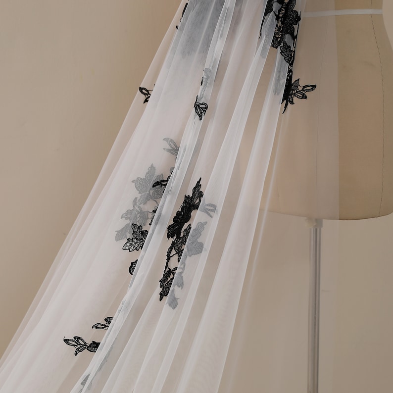 May include: White tulle fabric with black floral embroidery. The fabric is draped over a dress form.
