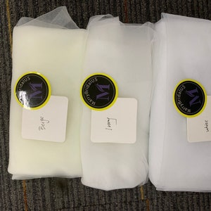 May include: Three white tulle fabric swatches with labels. The labels are yellow with black text and a purple circle with a white "M" inside. The labels read "Beige", "Ivory", and "White".