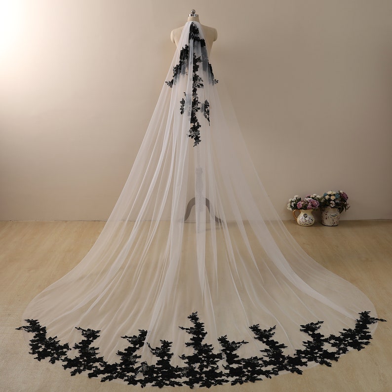 May include: A white tulle wedding veil with black floral lace trim. The veil is long and flowing, with the lace trim extending around the bottom edge.
