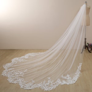 Peut inclure: Un long voile de mari&eacute;e en tulle blanc avec une d&eacute;licate bordure en dentelle florale. Le voile est drap&eacute; sur un mannequin et a un aspect fluide et &eacute;th&eacute;r&eacute;.