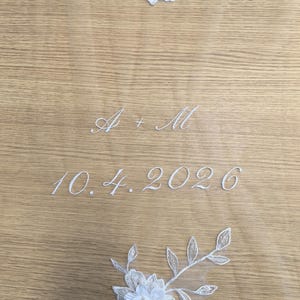 Może przedstawiać: Przezroczysty biały welon z białymi koronkowymi aplikacjami kwiatowymi na górze i na dole. Środek jest wyhaftowany białym kursywnym tekstem A + M, po którym następuje data 10.4.2026 w pasującej białej czcionce pisanej.