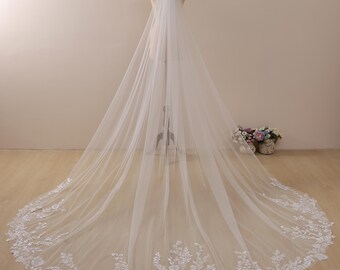 May include: A long white tulle wedding veil with a delicate floral lace trim. The veil is draped over a mannequin and has a cathedral length.