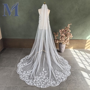May include: A long, white bridal veil with intricate floral embroidery along the scalloped edge. The veil is displayed on a mannequin, showcasing its delicate, flowing design. The veil's sheer fabric and detailed embellishments create an elegant, romantic aesthetic.
