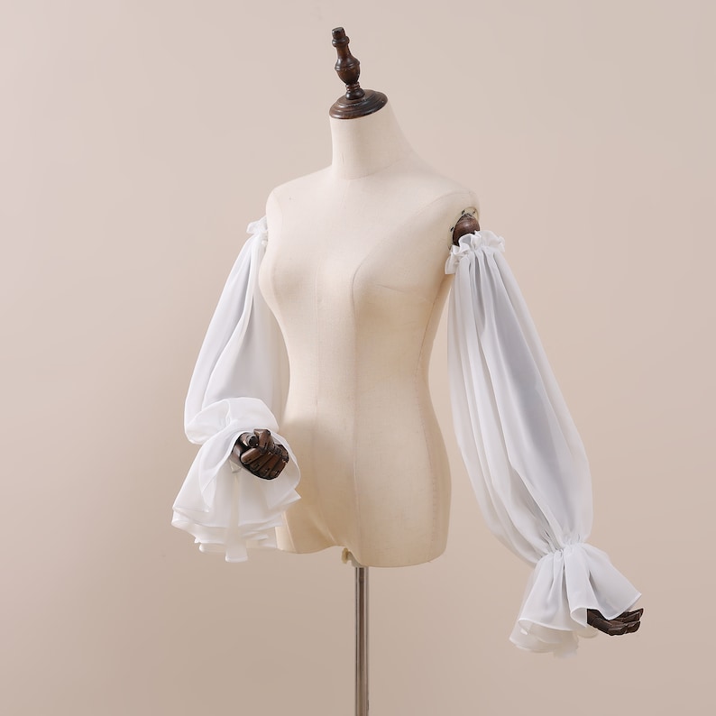 Pu&ograve; includere: Maniche bianche a spalle scoperte con polsini arricciati e tessuto voluminoso e trasparente. Le maniche sono esposte su un manichino, mostrando la silhouette fluida del design. L'indumento &egrave; probabilmente per un costume o un'occasione speciale.