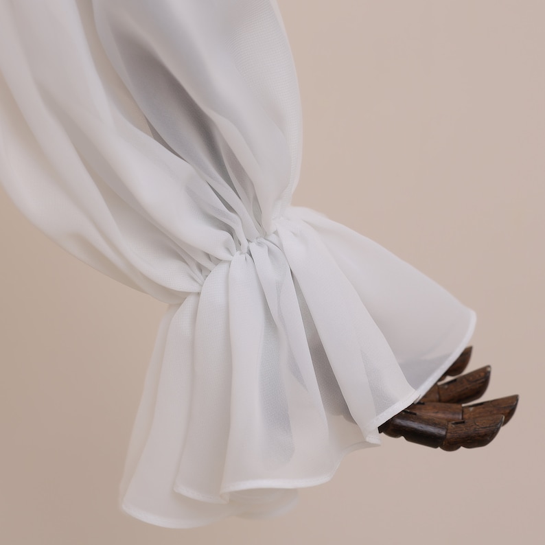 Pu&ograve; includere: Una manica in tessuto bianco e trasparente con polsino arricciato e bordo svasato e arruffato. La manica &egrave; drappeggiata su una forma di legno, mettendo in risalto la delicata trama e il design del capo.