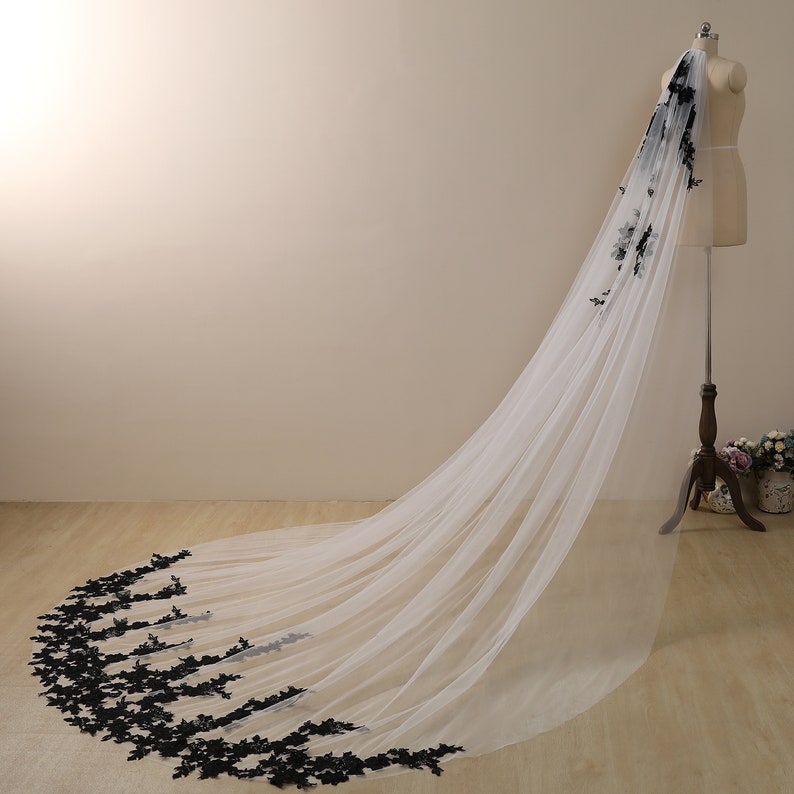 May include: A long white tulle wedding veil with black floral lace trim. The veil is draped over a dress form.