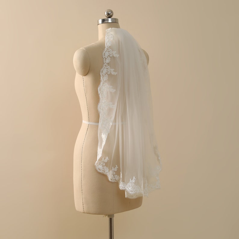 May include: A white bridal veil with a delicate lace trim. The veil is draped over a dress form.