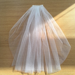 May include: A white bridal veil made of sheer fabric, with a delicate, beaded trim along the top edge. The veil is spread out, showcasing its flowing, layered design. The background is a light brown wooden surface.