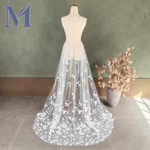 May include: A white tulle skirt with floral embroidery is displayed on a mannequin. The skirt is long, flowing, and features delicate white flowers. The letter "M" is visible in the upper left corner.